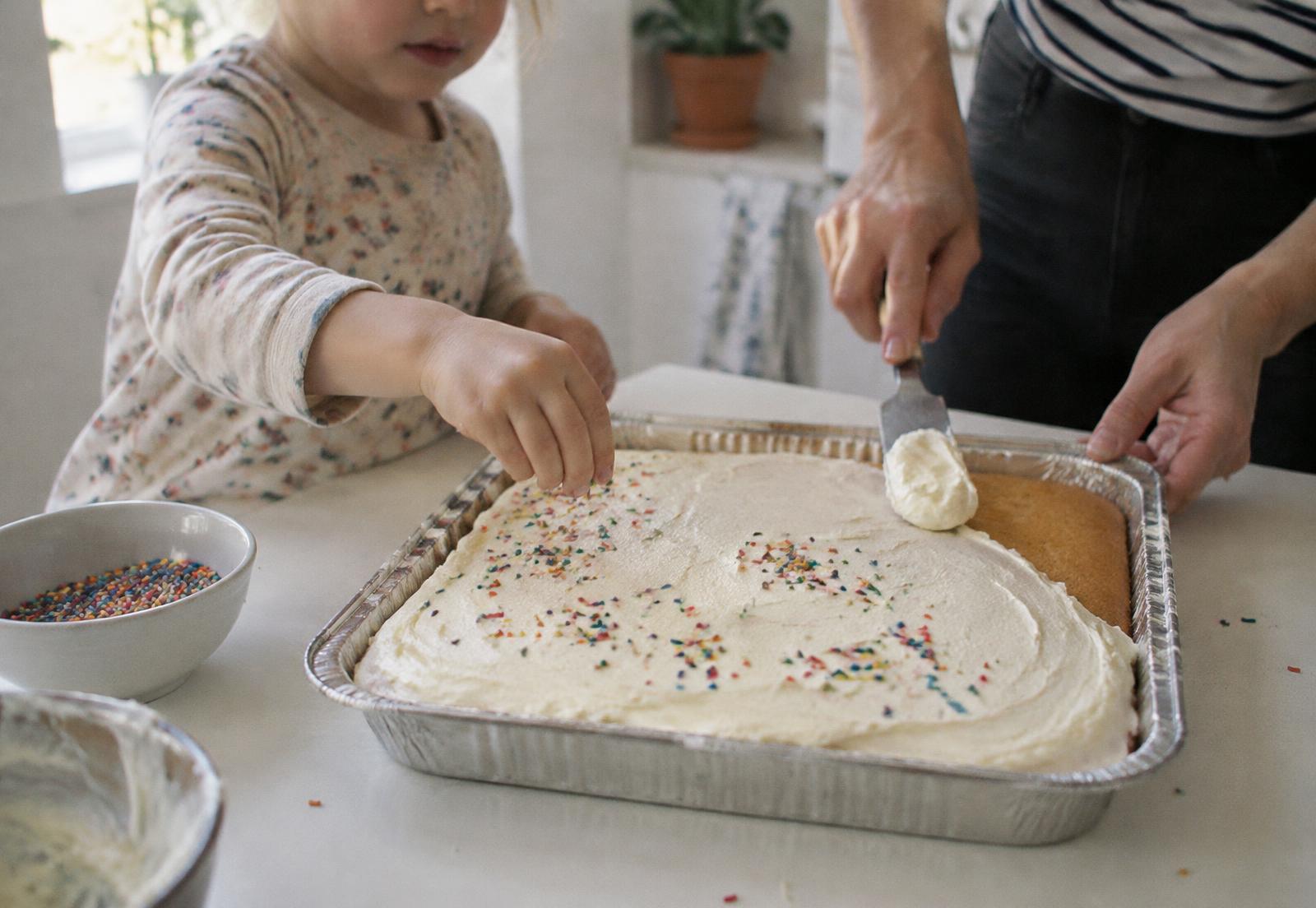 A parent and child frosting a homemade sheet cake in an aluminum pan